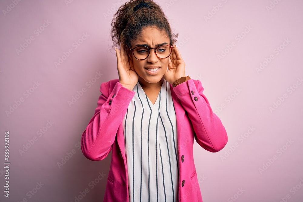 Beautiful african american businesswoman wearing jacket and glasses over pink background Trying to hear both hands on ear gesture, curious for gossip. Hearing problem, deaf
