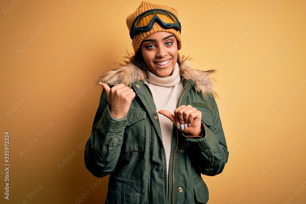 Young african american afro skier girl wearing snow sportswear and ski goggles Pointing to the back behind with hand and thumbs up, smiling confident