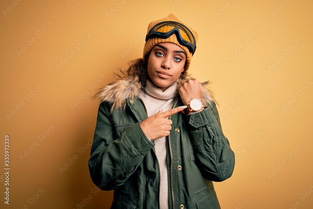 Young african american afro skier girl wearing snow sportswear and ski goggles In hurry pointing to watch time, impatience, looking at the camera with relaxed expression Young african american afro skier girl wearing snow sportswear and ski goggles In hurry pointing to watch time, impatience, looking at the camera with relaxed expression