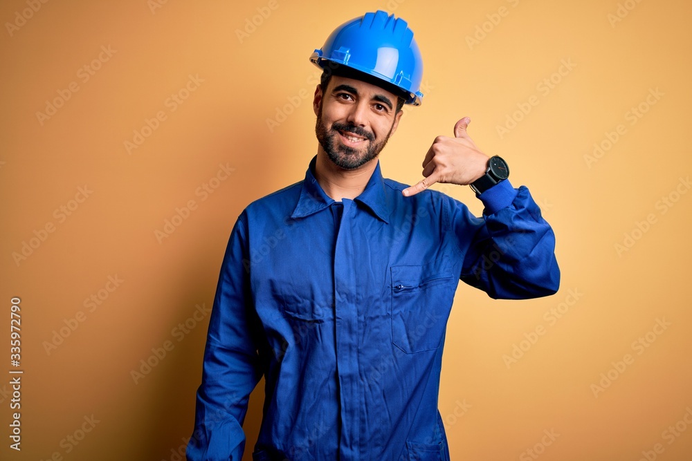 Mechanic man with beard wearing blue uniform and safety helmet over yellow background smiling doing phone gesture with hand and fingers like talking on the telephone. Communicating concepts.