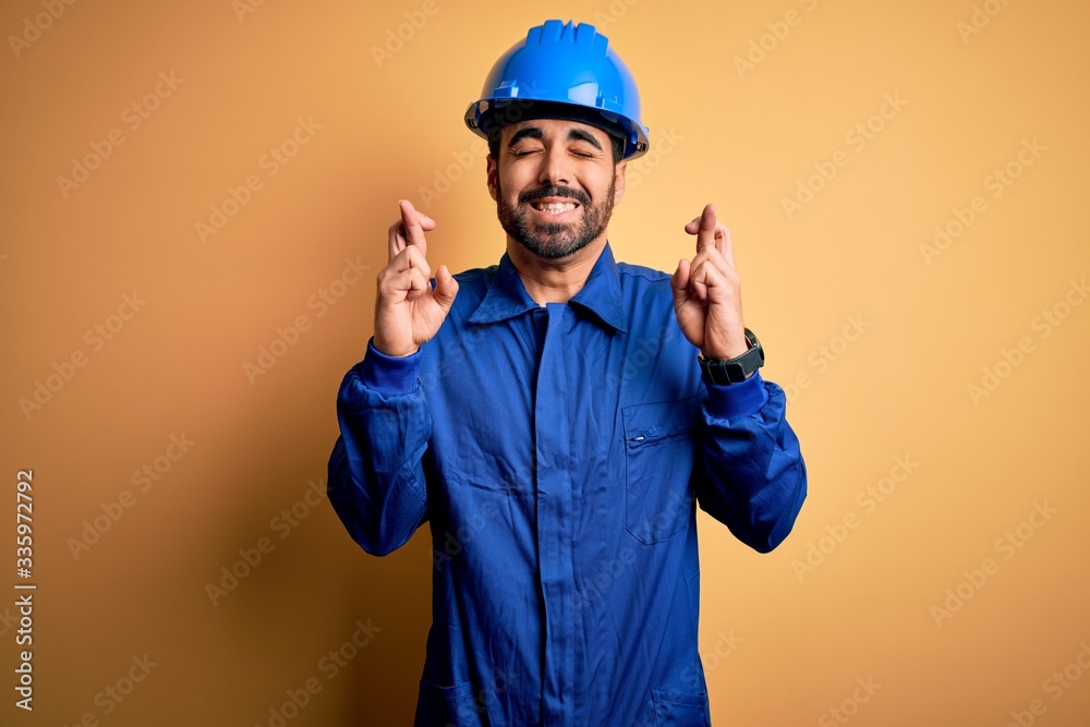 Mechanic man with beard wearing blue uniform and safety helmet over yellow background gesturing finger crossed smiling with hope and eyes closed. Luck and superstitious concept.