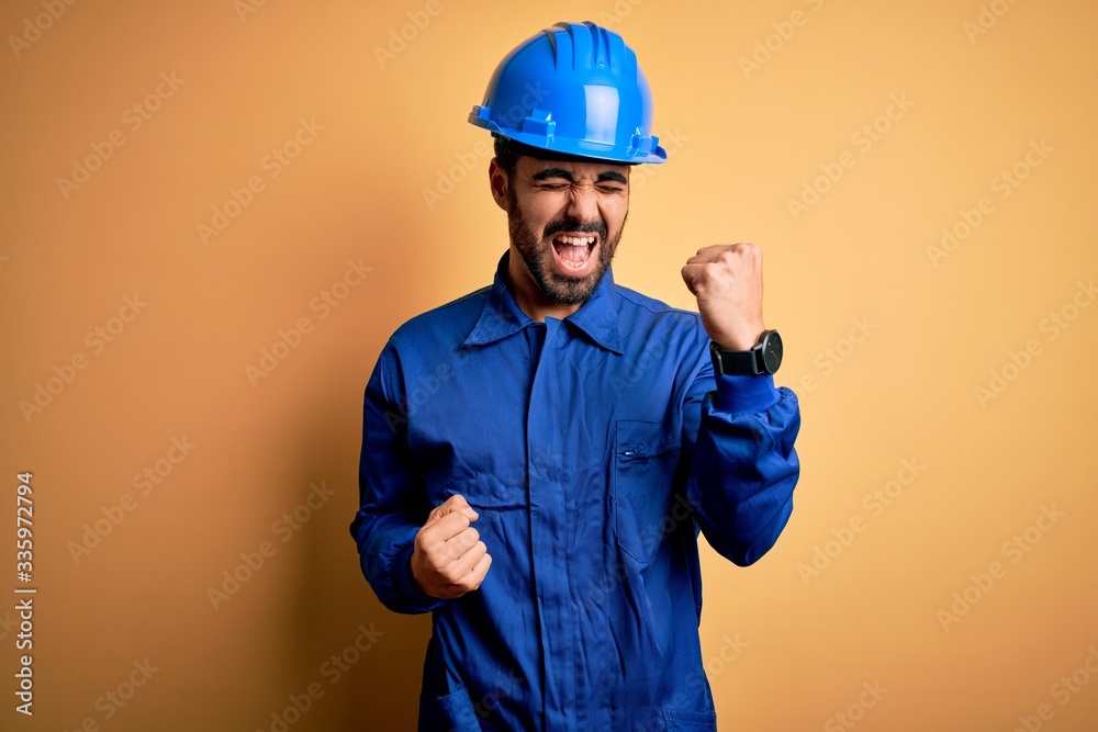 Mechanic man with beard wearing blue uniform and safety helmet over yellow background celebrating surprised and amazed for success with arms raised and eyes closed. Winner concept.