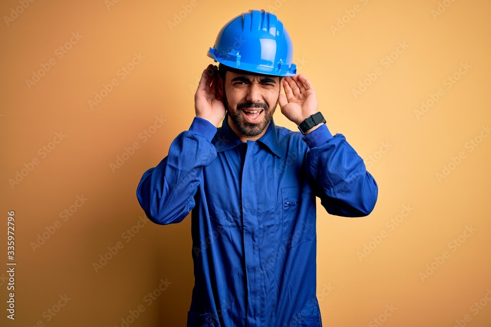 Mechanic man with beard wearing blue uniform and safety helmet over yellow background Trying to hear both hands on ear gesture, curious for gossip. Hearing problem, deaf Mechanic man with beard wearing blue uniform and safety helmet over yellow background Trying to hear both hands on ear gesture, curious for gossip. Hearing problem, deaf