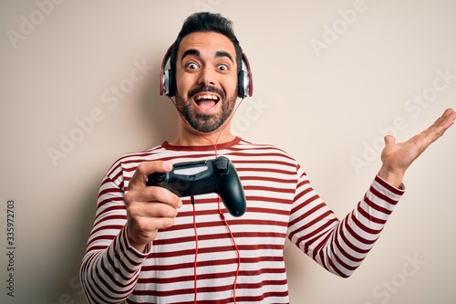 Young handsome gamer man with beard playing video game using joystick and headphones very happy and excited, winner expression celebrating victory screaming with big smile and raised hands