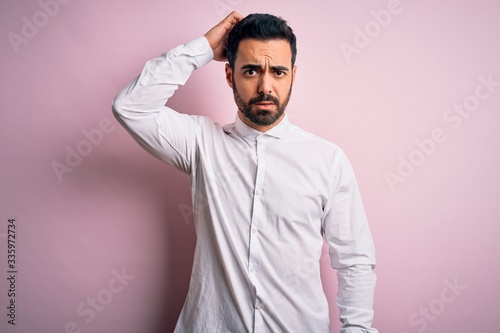 Young handsome man with beard wearing casual shirt standing over pink background confuse and wonder about question. Uncertain with doubt, thinking with hand on head. Pensive concept.