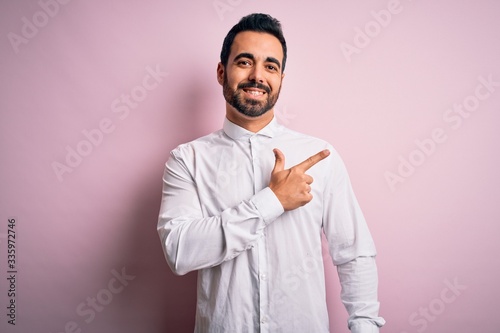 Young handsome man with beard wearing casual shirt standing over pink background cheerful with a smile of face pointing with hand and finger up to the side with happy and natural expression on face