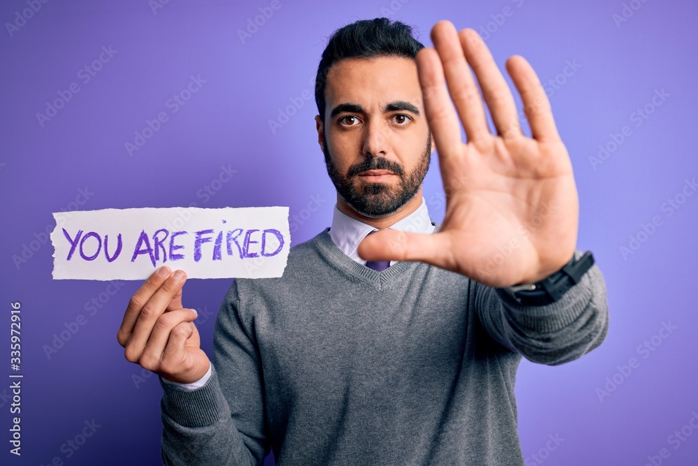 Young handsome businessman with beard holding paper with you are fired message with open hand doing stop sign with serious and confident expression, defense gesture Young handsome businessman with beard holding paper with you are fired message with open hand doing stop sign with serious and confident expression, defense gesture