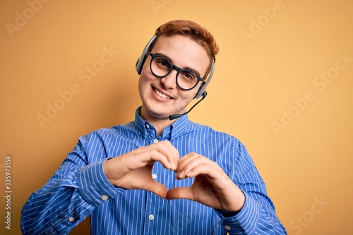 Young handsome redhead call center agent man wearing glasses working using headset smiling in love showing heart symbol and shape with hands. Romantic concept.