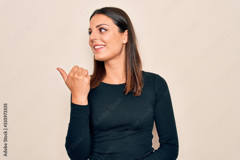 Young beautiful brunette woman wearing casual t-shirt standing over isolated white background smiling with happy face looking and pointing to the side with thumb up. Young beautiful brunette woman wearing casual t-shirt standing over isolated white background smiling with happy face looking and pointing to the side with thumb up.