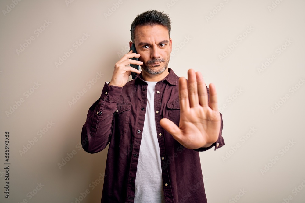 Young handsome man having conversation talking on the smartphone over white background with open hand doing stop sign with serious and confident expression, defense gesture Young handsome man having conversation talking on the smartphone over white background with open hand doing stop sign with serious and confident expression, defense gesture