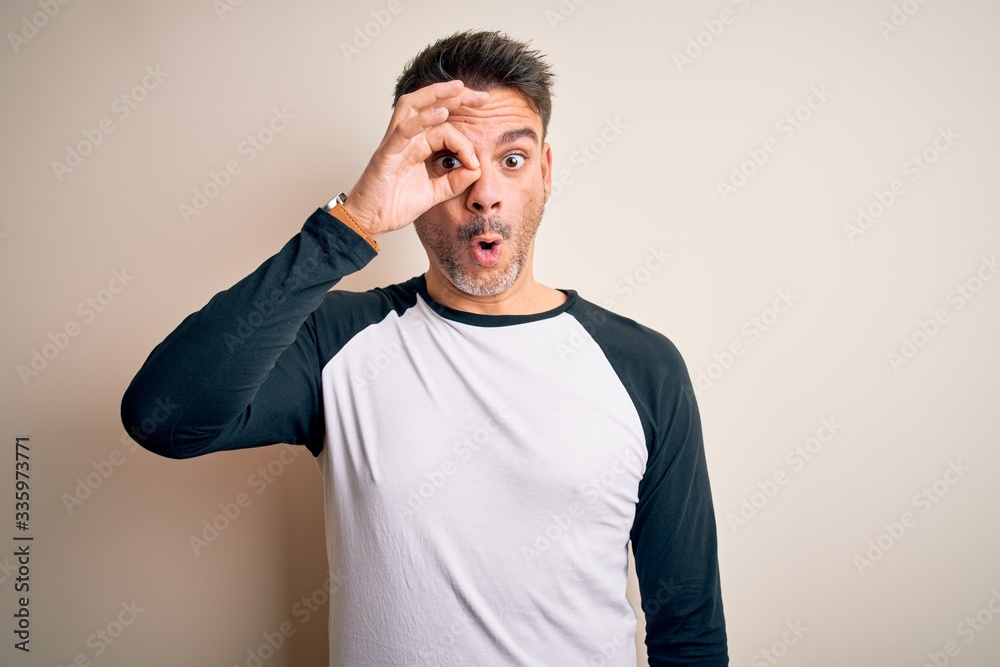 Young handsome man wearing casual t-shirt standing over isolated white background doing ok gesture shocked with surprised face, eye looking through fingers. Unbelieving expression. Young handsome man wearing casual t-shirt standing over isolated white background doing ok gesture shocked with surprised face, eye looking through fingers. Unbelieving expression.