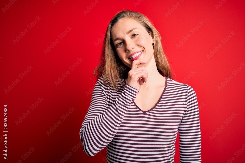 Young beautiful blonde woman wearing casual striped t-shirt over isolated red background looking confident at the camera with smile with crossed arms and hand raised on chin. Thinking positive. Young beautiful blonde woman wearing casual striped t-shirt over isolated red background looking confident at the camera with smile with crossed arms and hand raised on chin. Thinking positive.