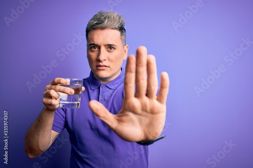 Young modern man drinking a glass of fresh water over purple isolated background with open hand doing stop sign with serious and confident expression, defense gesture