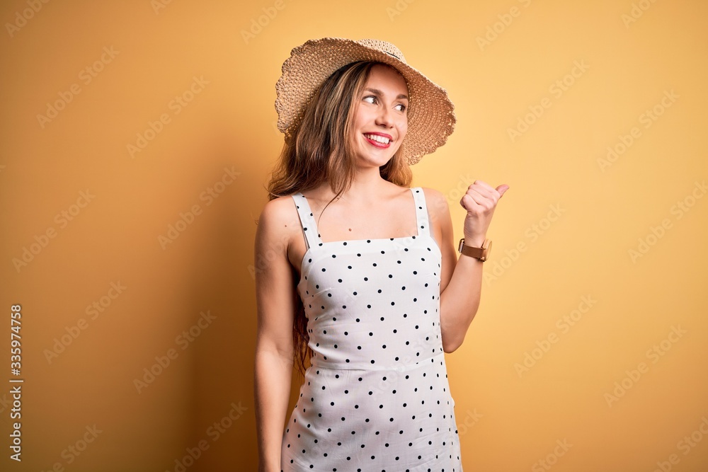 Young beautiful blonde woman on vacation wearing summer hat over yellow background smiling with happy face looking and pointing to the side with thumb up. Young beautiful blonde woman on vacation wearing summer hat over yellow background smiling with happy face looking and pointing to the side with thumb up.