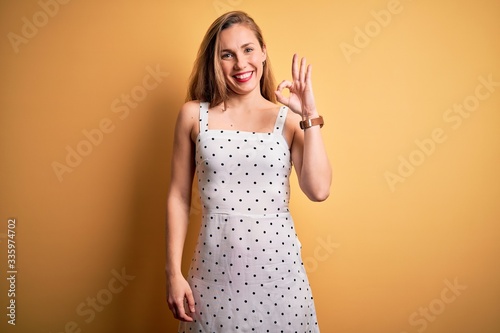 Young beautiful blonde woman on vacation wearing summer dress over yellow background smiling positive doing ok sign with hand and fingers. Successful expression.