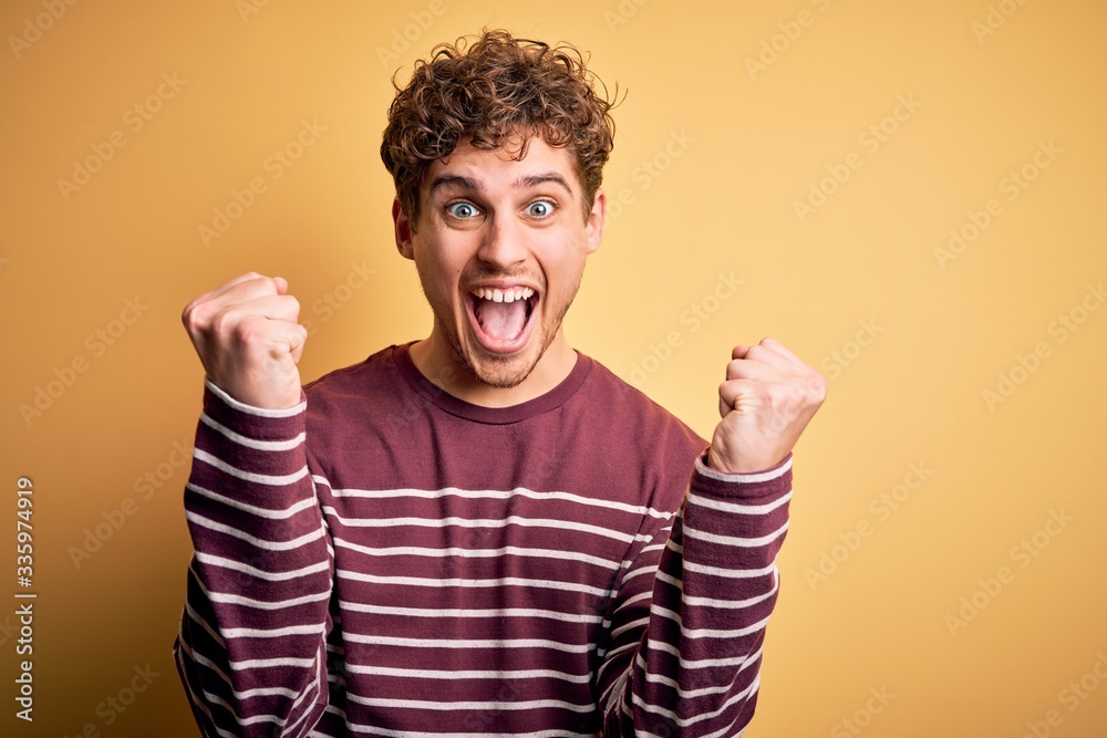 Young blond handsome man with curly hair wearing casual striped sweater celebrating surprised and amazed for success with arms raised and open eyes. Winner concept. Young blond handsome man with curly hair wearing casual striped sweater celebrating surprised and amazed for success with arms raised and open eyes. Winner concept.