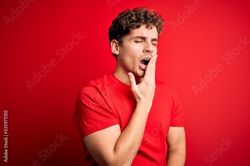 Young blond handsome man with curly hair wearing casual t-shirt over red background bored yawning tired covering mouth with hand. Restless and sleepiness. © Krakenimages.com