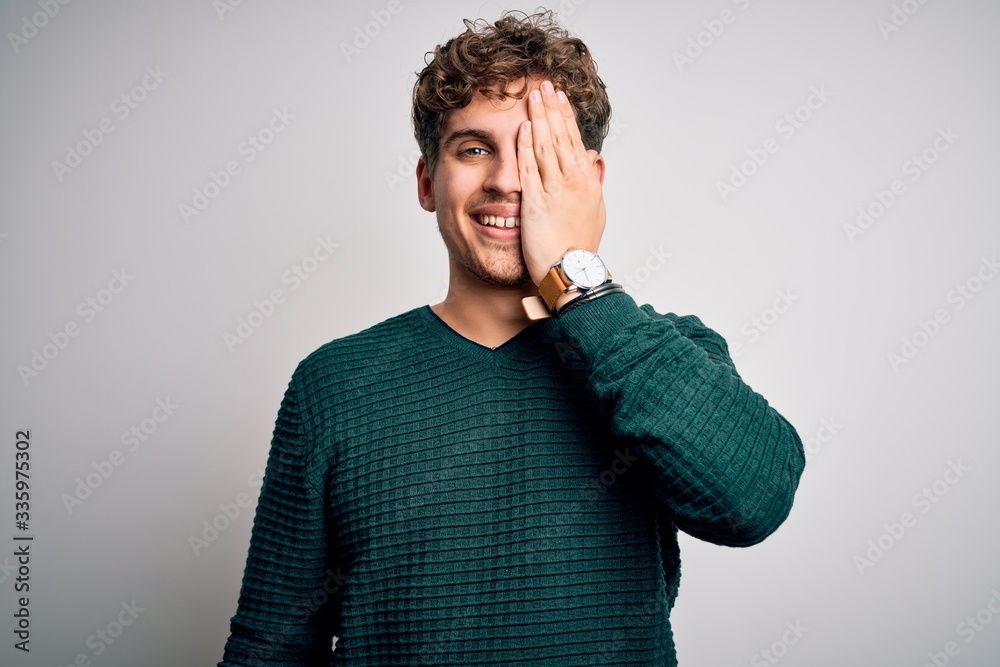 Young blond handsome man with curly hair wearing green sweater over white background covering one eye with hand, confident smile on face and surprise emotion. Young blond handsome man with curly hair wearing green sweater over white background covering one eye with hand, confident smile on face and surprise emotion.