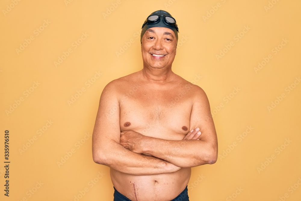 Middle age senior grey-haired swimmer man wearing swimsuit, cap and goggles happy face smiling with crossed arms looking at the camera. Positive person.