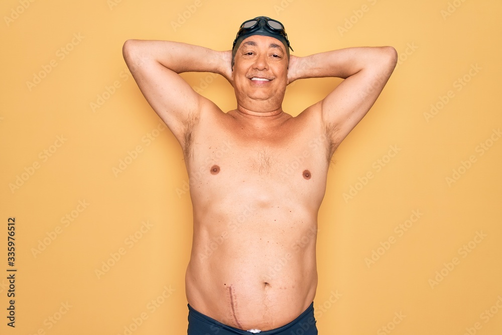 Middle age senior grey-haired swimmer man wearing swimsuit, cap and goggles relaxing and stretching, arms and hands behind head and neck smiling happy