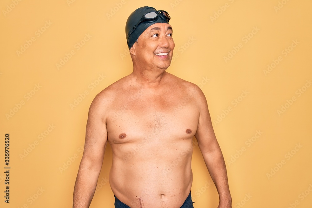 Middle age senior grey-haired swimmer man wearing swimsuit, cap and goggles looking away to side with smile on face, natural expression. Laughing confident.