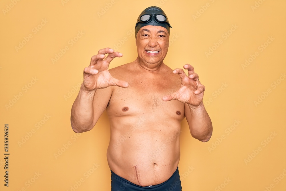 Middle age senior grey-haired swimmer man wearing swimsuit, cap and goggles smiling funny doing claw gesture as cat, aggressive and sexy expression