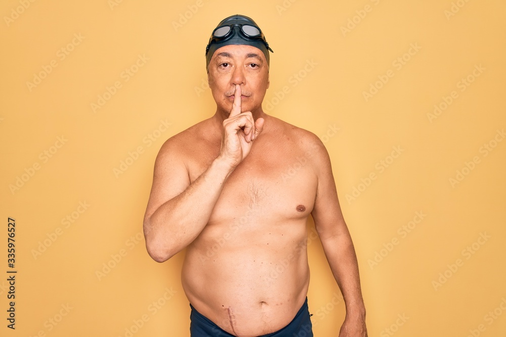 Middle age senior grey-haired swimmer man wearing swimsuit, cap and goggles asking to be quiet with finger on lips. Silence and secret concept.