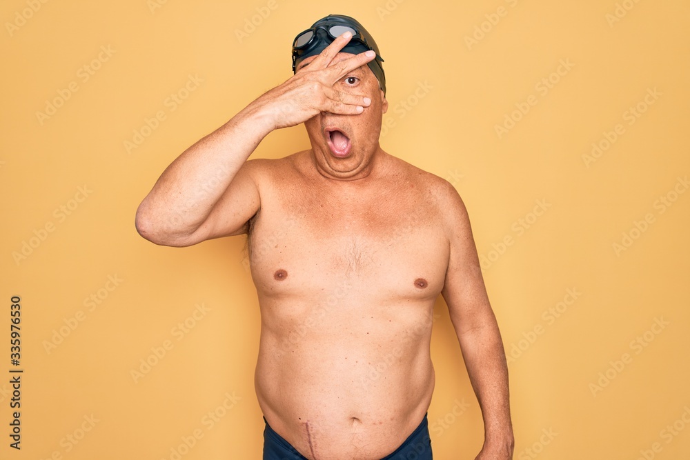 Middle age senior grey-haired swimmer man wearing swimsuit, cap and goggles peeking in shock covering face and eyes with hand, looking through fingers with embarrassed expression.