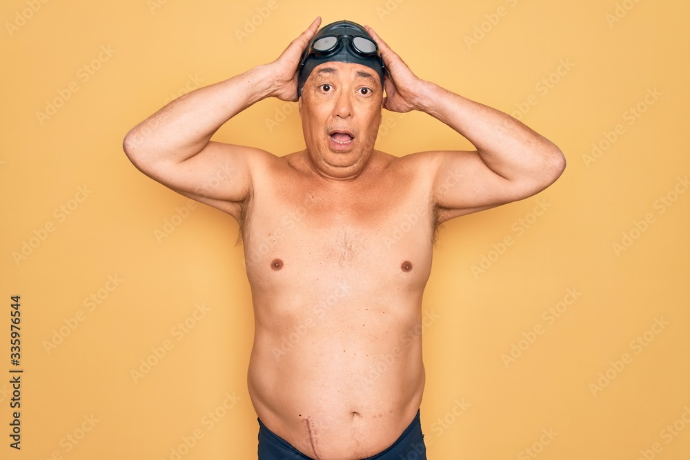 Middle age senior grey-haired swimmer man wearing swimsuit, cap and goggles Crazy and scared with hands on head, afraid and surprised of shock with open mouth