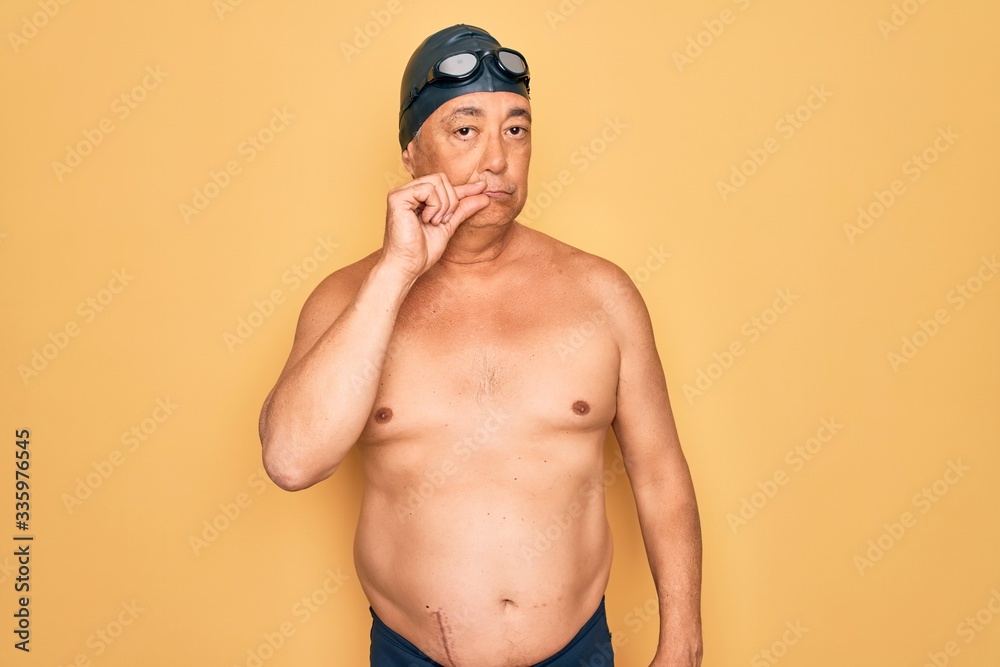 Middle age senior grey-haired swimmer man wearing swimsuit, cap and goggles mouth and lips shut as zip with fingers. Secret and silent, taboo talking