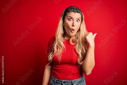 Young beautiful blonde woman wearing casual t-shirt standing over isolated red background Surprised pointing with hand finger to the side, open mouth amazed expression. © Krakenimages.com