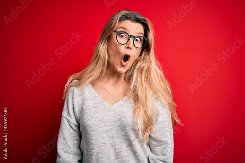 Young beautiful blonde woman wearing sweater and glasses over isolated red background afraid and shocked with surprise expression, fear and excited face. © Krakenimages.com