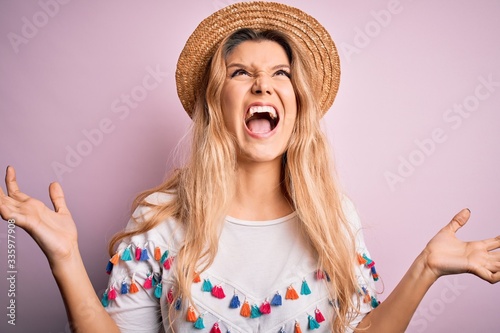 Young beautiful blonde woman wearing t-shirt and hat over isolated pink background crazy and mad shouting and yelling with aggressive expression and arms raised. Frustration concept.