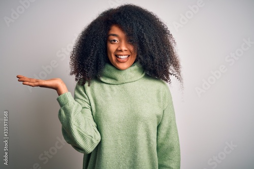 Young beautiful african american woman with afro hair wearing green winter sweater smiling cheerful presenting and pointing with palm of hand looking at the camera. © Krakenimages.com