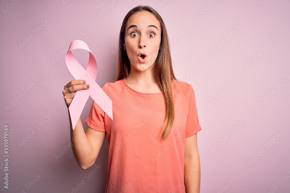 Young beautiful woman holding pink cancer ribbon standing over isolated pink background scared in shock with a surprise face, afraid and excited with fear expression Young beautiful woman holding pink cancer ribbon standing over isolated pink background scared in shock with a surprise face, afraid and excited with fear expression