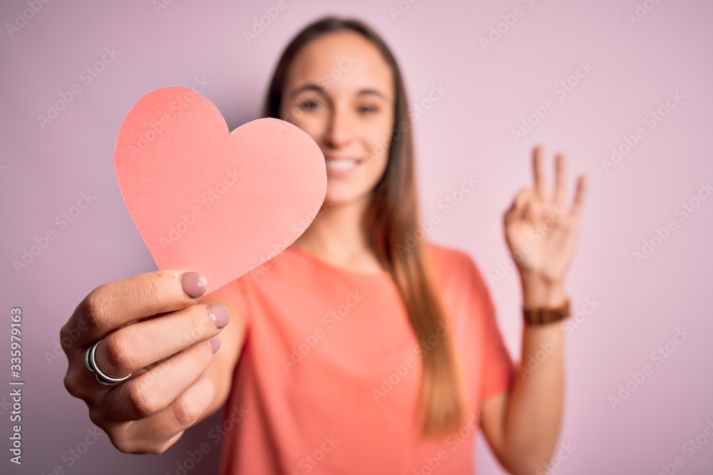 Young beautiful romantic woman holding paper heart shape over isolated pink background doing ok sign with fingers, excellent symbol Young beautiful romantic woman holding paper heart shape over isolated pink background doing ok sign with fingers, excellent symbol