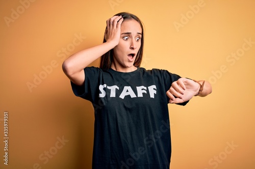 Young beautiful worker woman wearing staff uniform t-shirt over isolated yellow background Looking at the watch time worried, afraid of getting late