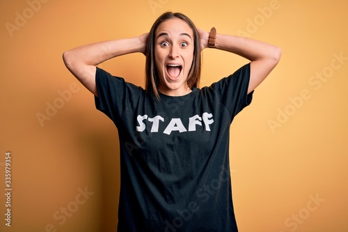 Young beautiful worker woman wearing staff uniform t-shirt over isolated yellow background Crazy and scared with hands on head, afraid and surprised of shock with open mouth photo