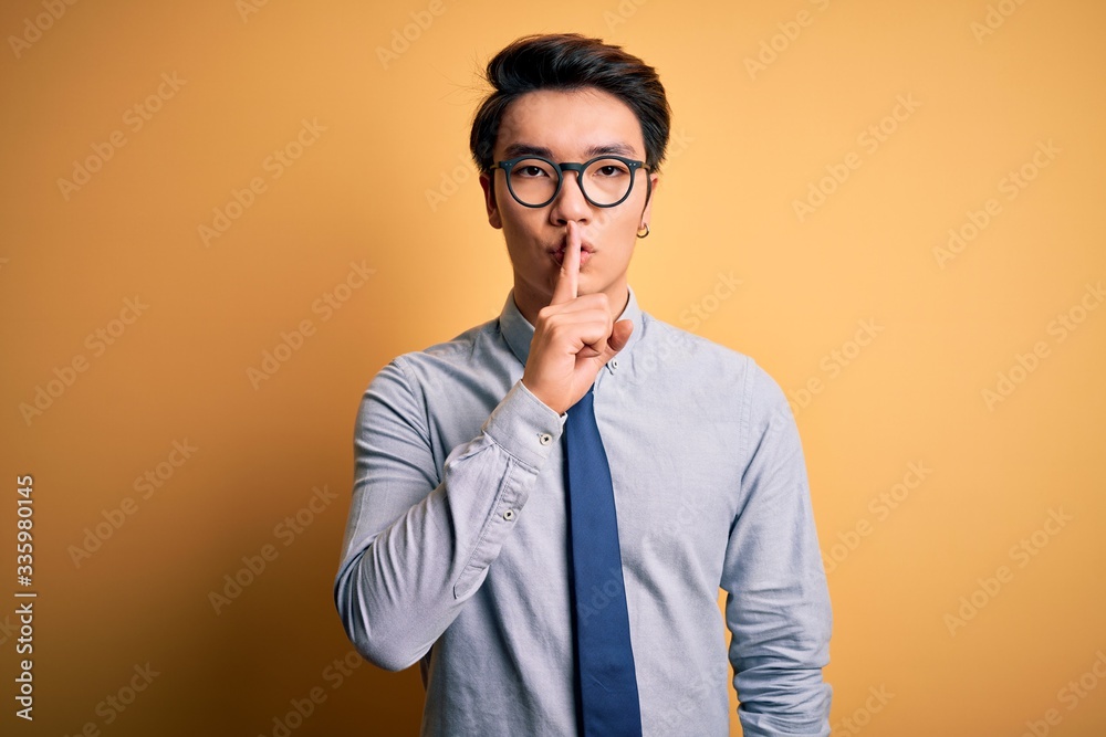 Young handsome chinese businessman wearing glasses and tie over yellow background asking to be quiet with finger on lips. Silence and secret concept. Young handsome chinese businessman wearing glasses and tie over yellow background asking to be quiet with finger on lips. Silence and secret concept.