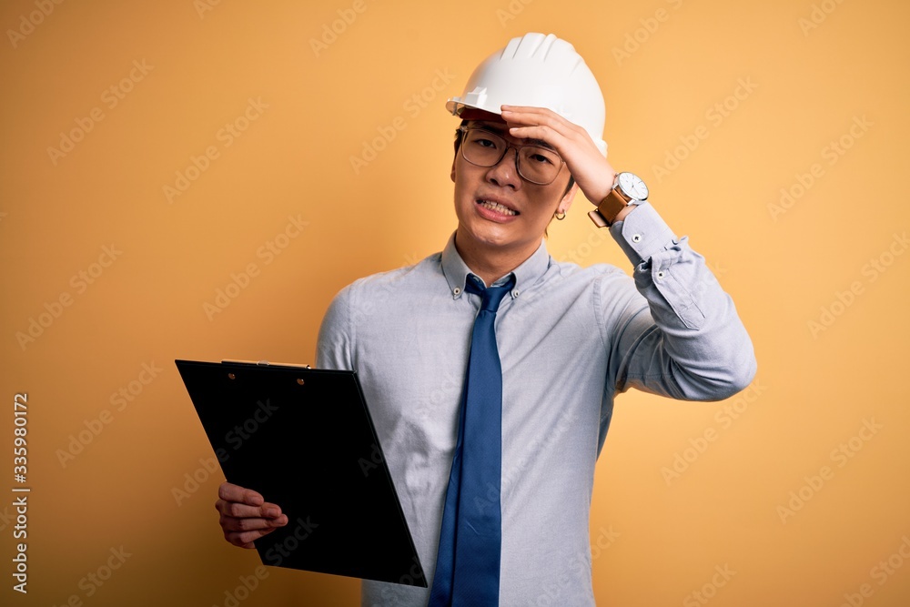 Young handsome chinese engineer man wearing safety helmet holding clipboard stressed with hand on head, shocked with shame and surprise face, angry and frustrated. Fear and upset for mistake. Young handsome chinese engineer man wearing safety helmet holding clipboard stressed with hand on head, shocked with shame and surprise face, angry and frustrated. Fear and upset for mistake.