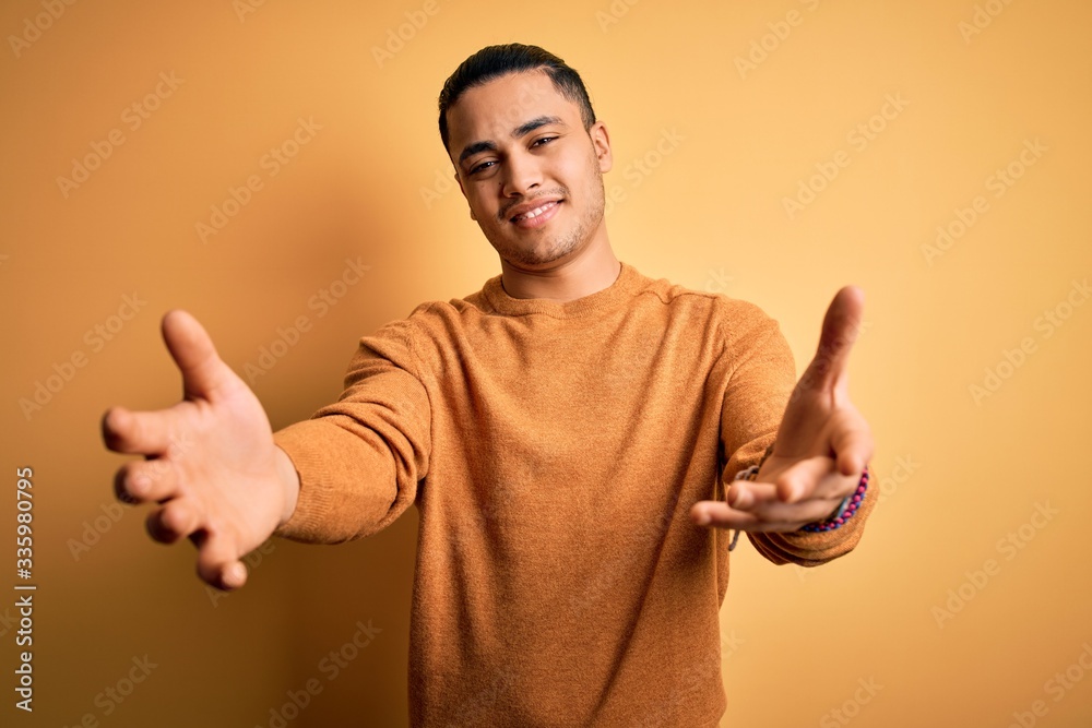 Young brazilian man wearing casual sweater standing over isolated yellow background looking at the camera smiling with open arms for hug. Cheerful expression embracing happiness.