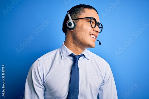 Young brazilian call center agent man wearing glasses and tie working using headset looking away to side with smile on face, natural expression. Laughing confident.