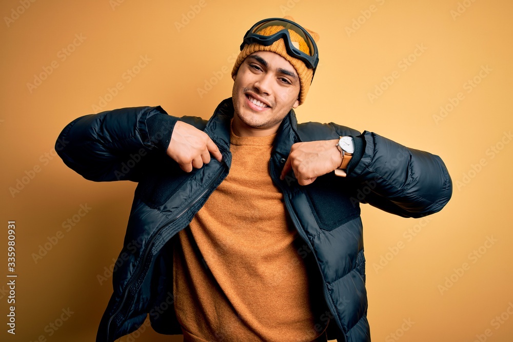 Young brazilian skier man wearing snow sportswear and ski goggles over yellow background looking confident with smile on face, pointing oneself with fingers proud and happy. Young brazilian skier man wearing snow sportswear and ski goggles over yellow background looking confident with smile on face, pointing oneself with fingers proud and happy.