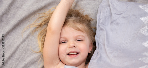 Portrait of a blue-eyed little girl with blond curly hair waking up in the morning on a gray bed laughing merrily.