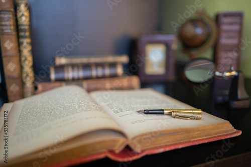An Antique golden fountain pen, on an old book.  Magnifying glass, clock and more antique books in the background © LaSu