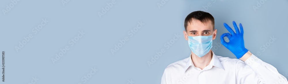A man in a medical mask shows the OK gesture in a blue medical glove, wearing a white shirt on a gray background, left empty space for text. Banner. Coronavirus, Covid-19 A man in a medical mask shows the OK gesture in a blue medical glove, wearing a white shirt on a gray background, left empty space for text. Banner. Coronavirus, Covid-19