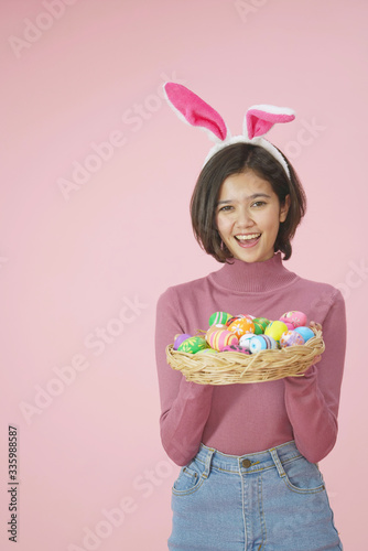 Cute girl smilling with bunny ears and basket full of Easter eggs on pink background