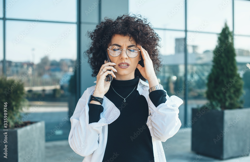 Thoughtful caucasian lady with curly hair and glasses touching her hair and posing outside during a business phone talk Thoughtful caucasian lady with curly hair and glasses touching her hair and posing outside during a business phone talk