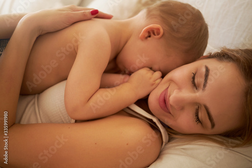 Close up shot of happy charming young mother lying on bed with her sleepy little child on chest. Pretty mom and infant cuddling in bedroom. Love, happiness, maternity and parenting concept