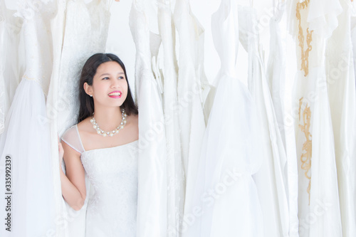 A beautiful Asian woman, Thai, is wearing a wedding dress standing in the midst of many white brides in the dress rental shop or boutique in the morning.